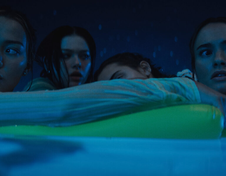 Four young women, partially submerged in water at night, cling to a yellow pool float. Their expressions are tense and concerned as they look in different directions. The scene is dimly lit with blue tones.
