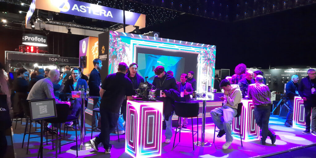 People gather and talk around a brightly lit stand with neon lights at a technology or trade show. Some are seated, some are standing, and multiple branded displays and equipment are visible in the background.