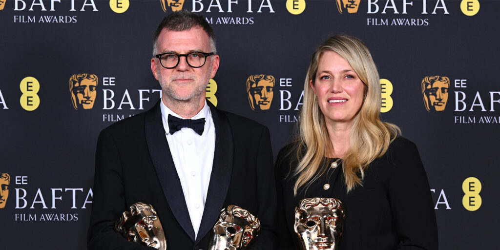 Two people stand in front of a BAFTA Film Awards backdrop, each holding a BAFTA trophy. The man wears a black tuxedo with glasses and a bow tie, whilst the woman wears a black outfit and has long blonde hair.