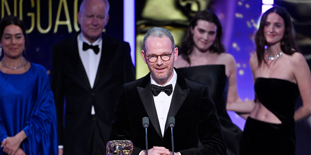 A man in a black dinner jacket and glasses speaks at a lectern with microphones, holding a BAFTA award. Four people, two men and two women in formal attire, stand behind him on stage under bright lighting.