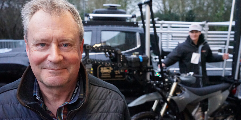 A man in a dark jacket stands in the foreground, smiling at the camera during the BSC Expo 2026. In the background, another person stands near a camera rig, a motorbike, and a large black vehicle, with trees visible behind them.