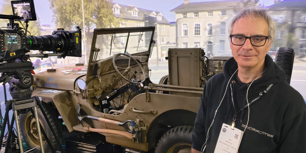 A man wearing glasses and a black hoodie stands beside a vintage military jeep at BSC Expo 2026, with a film camera set-up to the left. The background shows a blurred image of buildings and streets, capturing the event review atmosphere.