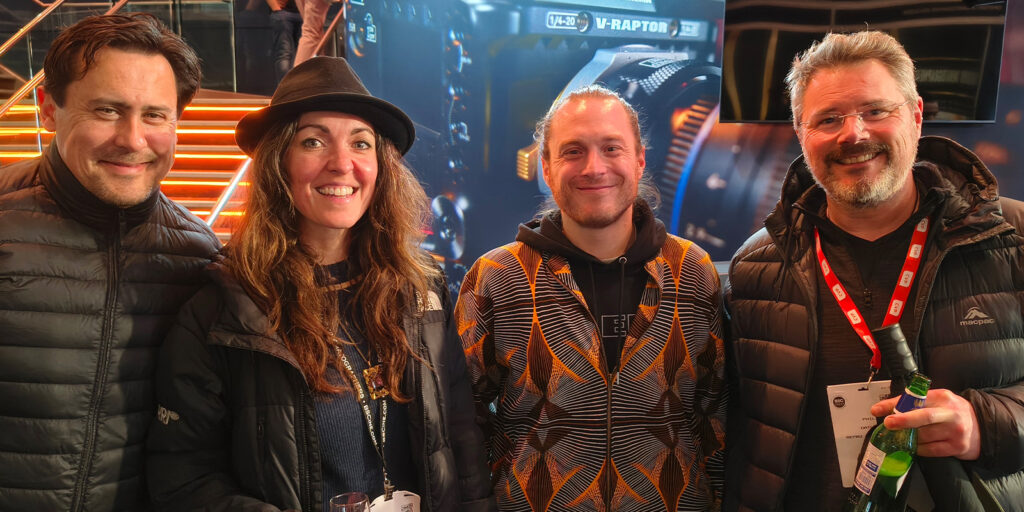 Four people stand indoors at the BSC Expo 2026, smiling at the camera in casual, warm clothing. One person holds a drink, and two wear lanyards with badges. Orange-lit stairs and a screen set the backdrop for this lively event review moment.