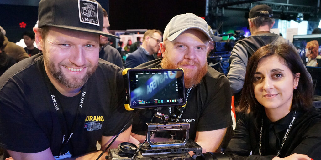 Three people pose behind a camera rig at an event. The two men wear hats and lanyards, whilst the woman on the right has shoulder-length dark hair. There are people and equipment in the background.