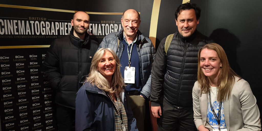 Five people, three men and two women, stand and sit in front of a wall with British Cinematographer repeated on it. They are wearing jackets and have lanyards, suggesting they are at an event or conference.