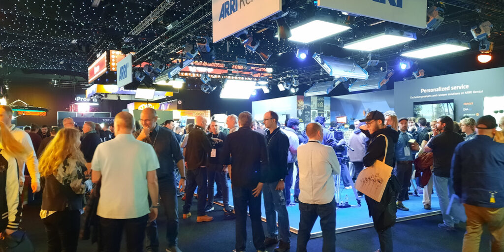 A crowd of people stands and walks around an indoor exhibition or trade show with various stands, illuminated signs, and displays under a ceiling filled with small lights.