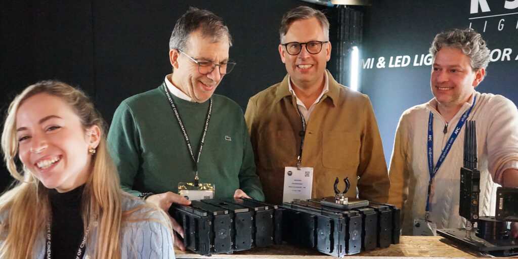 Four people stand behind a table displaying stacked electronic devices at BSC Expo 2026. Three men in the background smile with conference badges, whilst a woman in the foreground smiles towards the camera, capturing a lively event review atmosphere.