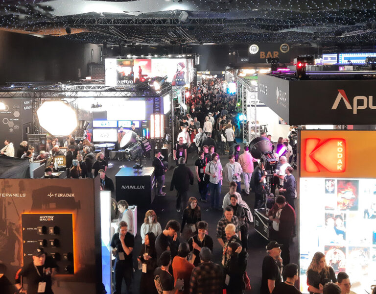 A crowded convention hall with numerous stands displaying technology and media equipment, large illuminated brand signs such as Aputure and Kodak, and many people walking and interacting with exhibitors.