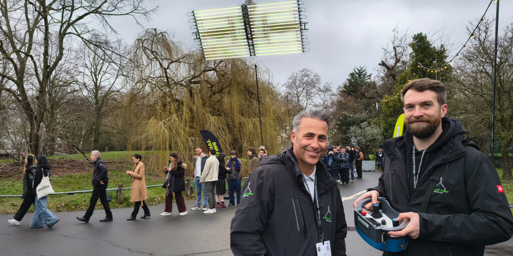 Two men in black jackets stand in the foreground, one holding a remote control. Behind them, people queue along a park pathway with trees. Bright tube lights are suspended above at the BSC Expo 2026, and the sky is overcast.