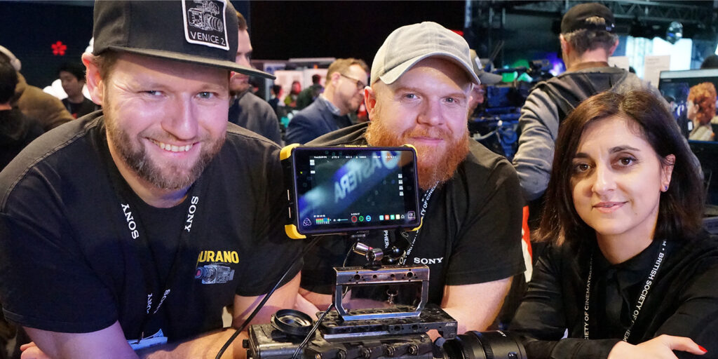 Three people are gathered around a professional video camera rig with an external monitor at the bustling BSC Expo. Two men are on the left, and a woman is on the right. All are facing the camera and smiling in this vibrant event review setting.