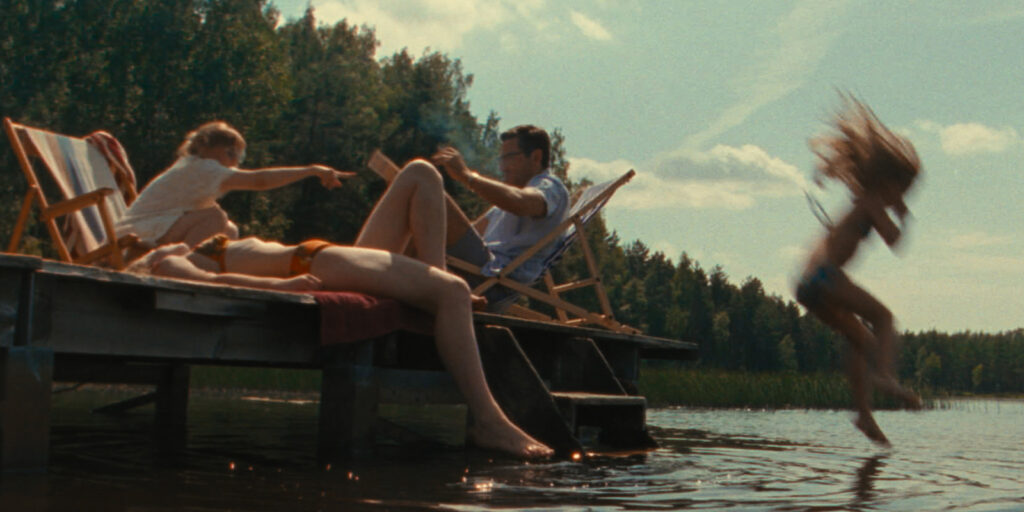 Three people relax on a wooden jetty by a lake; one person reclines on a lounger, another sits upright, and a third person leaps into the water. Trees and blue sky are visible in the background.