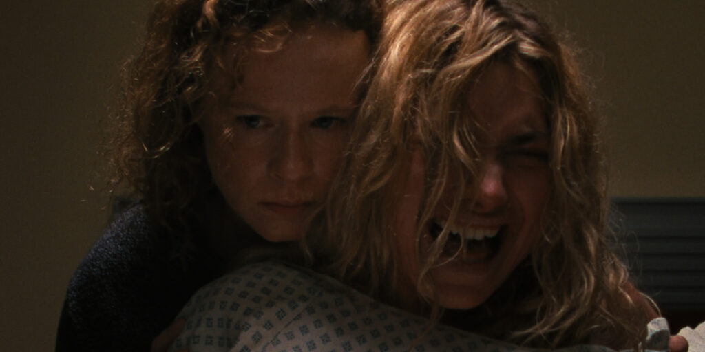 Two women are in a dimly lit room; one, wearing a hospital gown, appears distressed and is screaming, while the other, behind her, looks forward with a tense, serious expression.