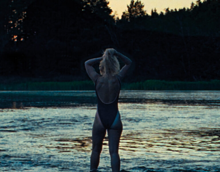 A person in a dark swimming costume stands in shallow water at dusk, facing away and tying up their hair, with trees silhouetted in the background.