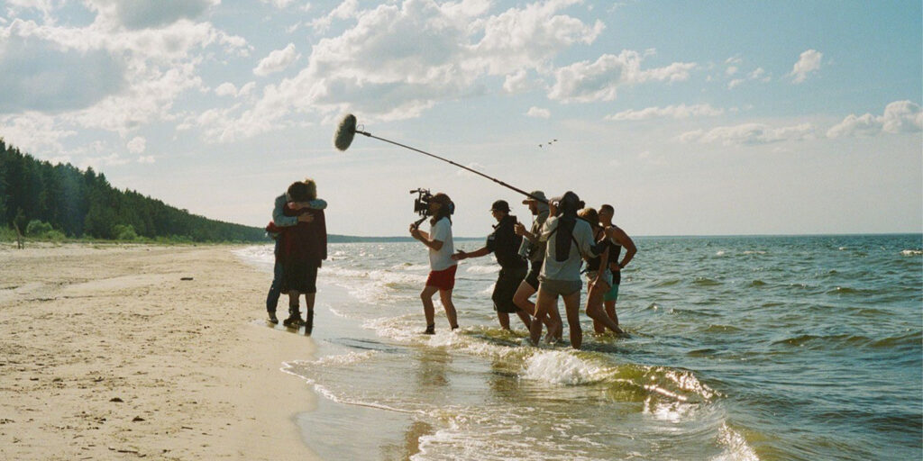 A film crew records two people hugging on a sandy beach. Crew members hold a boom microphone, camera, and other equipment while standing in shallow water near the shore under a partly cloudy sky.