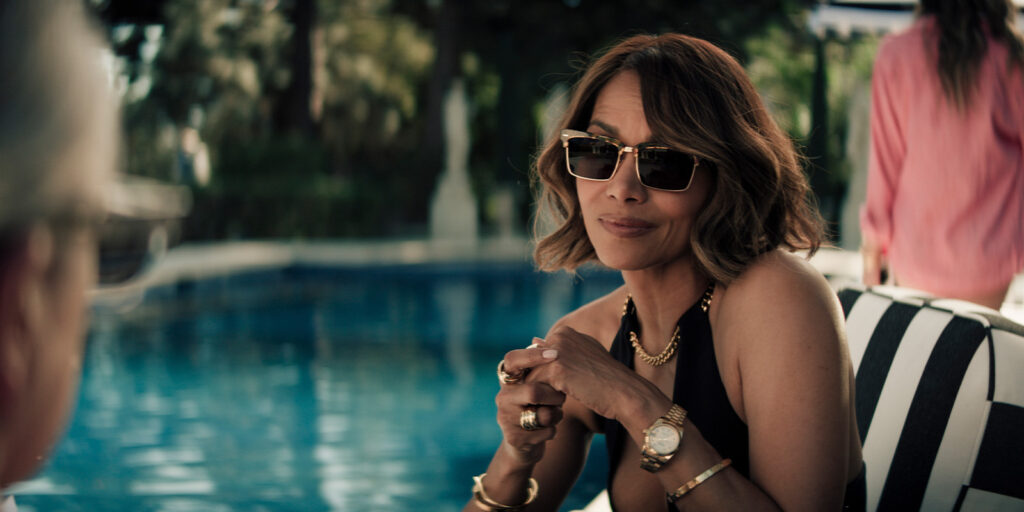 A woman wearing sunglasses, gold jewellery, and a black top sits by a swimming pool on a black and white striped chair, looking towards someone off-camera. The pool and greenery are visible in the background.