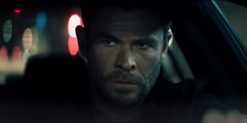 A man with a serious expression is sitting in a car at night, gripping the steering wheel. The background shows blurred red and blue lights.