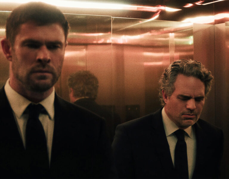Two men in suits stand in a lift with mirrored walls, both looking serious. The lighting is dim and warm, creating a reflective effect with multiple reflections of the men visible in the background.