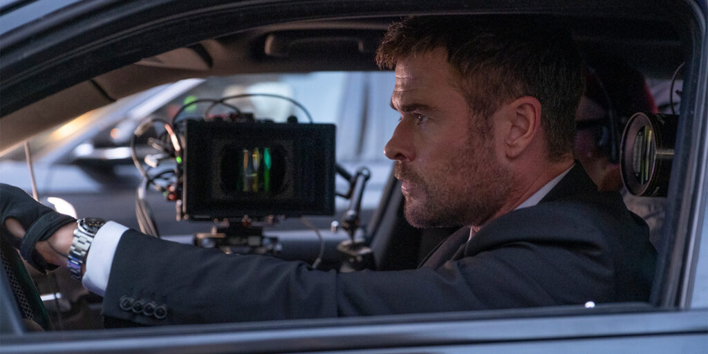 A man in a suit sits in the driver’s seat of a car, gripping the steering wheel. A film camera rig is attached inside the car, indicating a scene is being filmed. The man looks focused and serious.