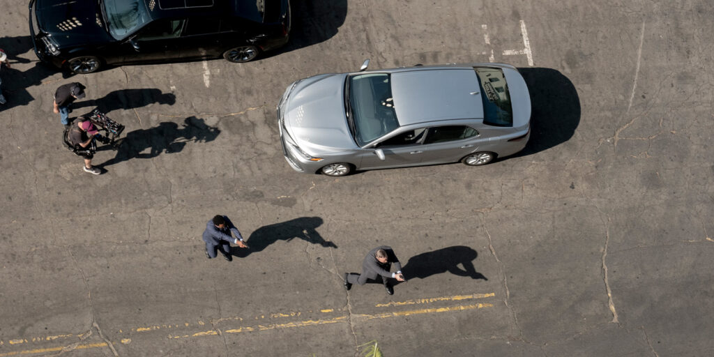 An overhead view of a silver car parked on a street with several people walking nearby. Faded yellow lines are visible on the tarmac, and part of a black car is parked to the left. Shadows are cast on the ground.
