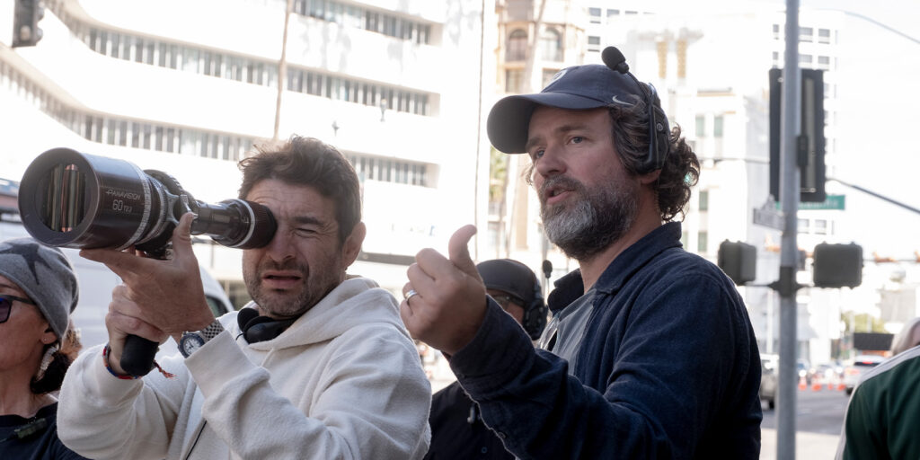 Two men stand outdoors; one looks through a film camera lens while the other speaks and gestures with his thumb up. Both wear casual clothing and headsets, with buildings and street signs visible in the background.