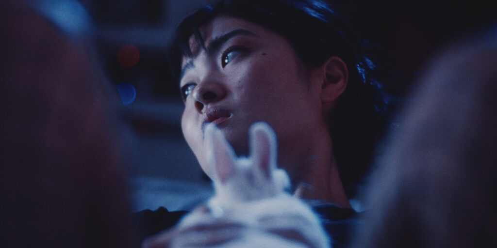 A woman with short dark hair looks to the side in a dimly lit setting with blue tones. In the foreground, a white rabbit is partially visible, its ears and head in focus.