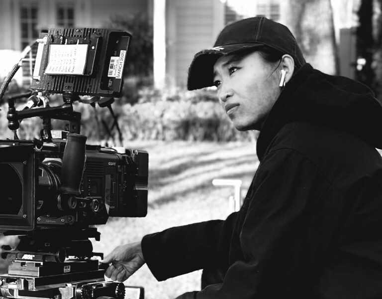 A person wearing a cap and hoodie sits behind a professional video camera on a film set outdoors, with another person in sunglasses partially visible in the background.