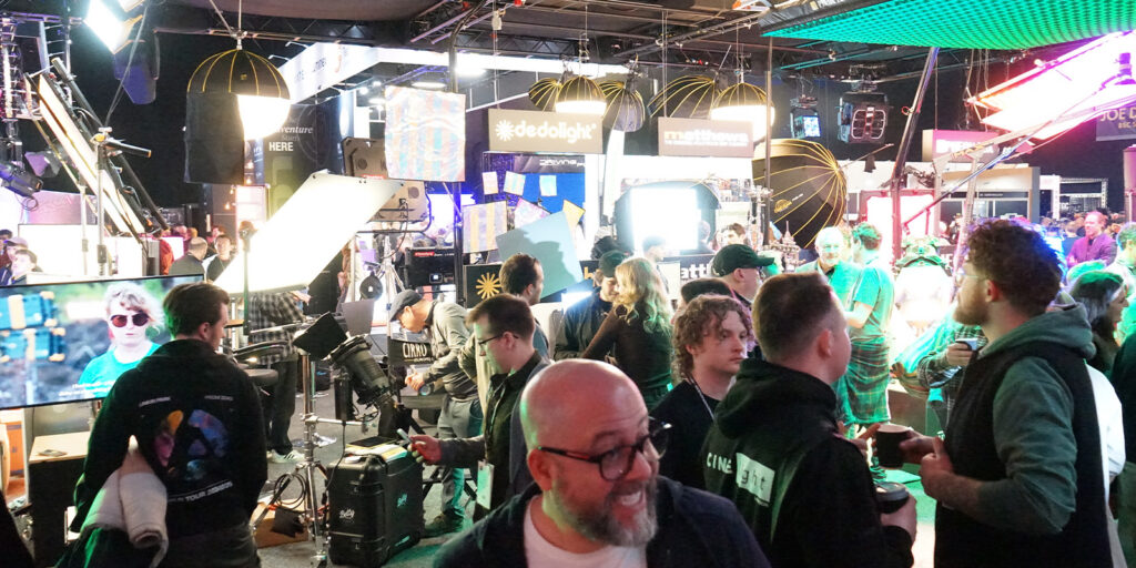 A crowded indoor event with people standing and chatting around exhibition stands, illuminated by bright lights and electronic displays, with various equipment and banners visible in the background.