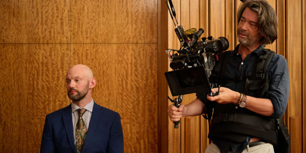 A man in a suit sits against a wooden wall while another man stands beside him operating a professional video camera mounted on a stabilising rig.