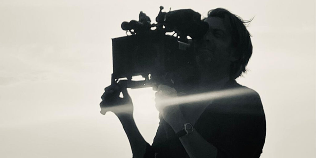 A person is holding a large professional video camera outdoors. The image is in black and white, and the person is silhouetted against a bright sky with sunlight shining through.