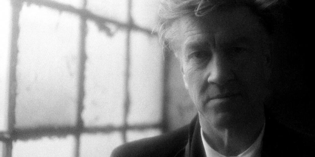 A black-and-white photo of a man in a suit, shown in soft focus. He stands indoors near a large window with frosted glass panes, with light streaming in from behind. His expression is neutral.
