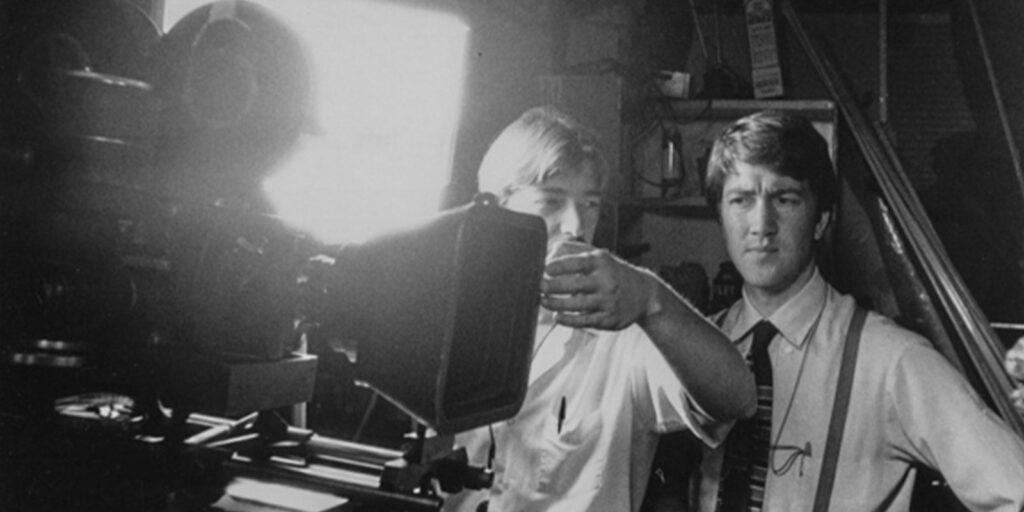 Two men stand next to a film camera, one adjusting the lens and the other watching intently. There is bright studio lighting behind them, suggesting a film set or production environment.