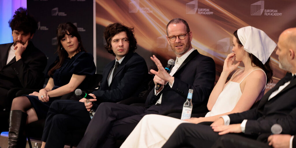 A group of six people in formal attire sit on stage in front of a backdrop, taking part in a panel discussion. One person is speaking and gesturing with his hand whilst holding a microphone.
