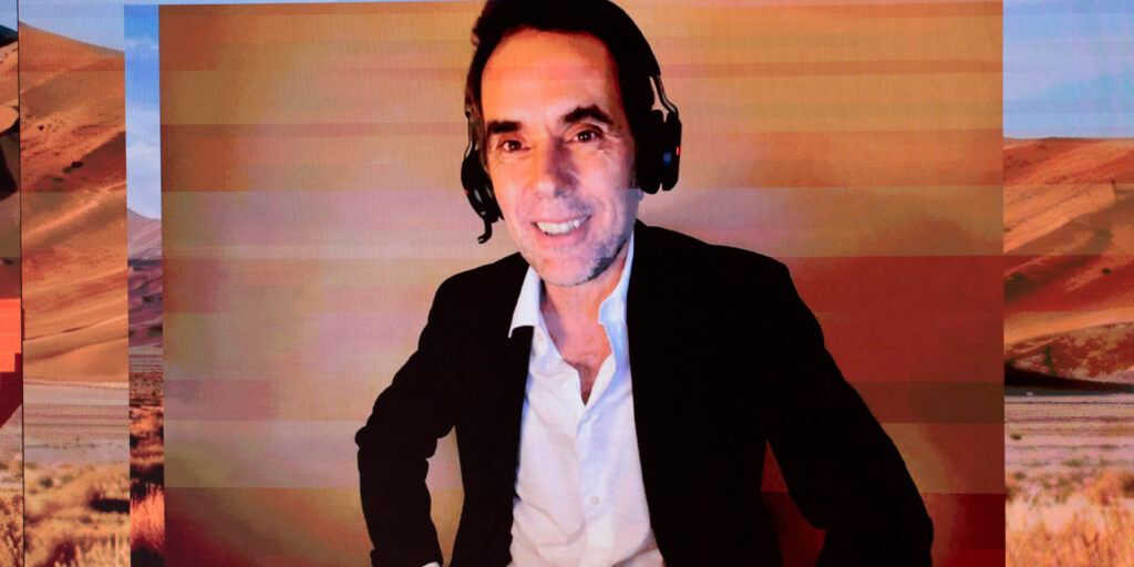 A man wearing a black blazer, white shirt, and headset smiles whilst sitting in front of a background showing a desert landscape with pixelation effects.