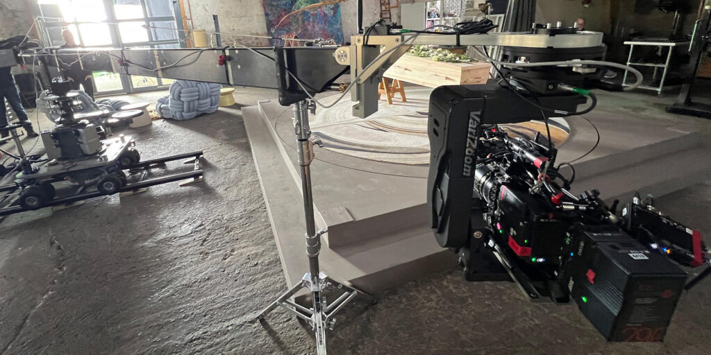 A professional film camera is mounted on a Varizoom camera crane inside an industrial studio. Nearby are other film equipment, including a camera dolly and lighting stands. The area has concrete floors and steps.