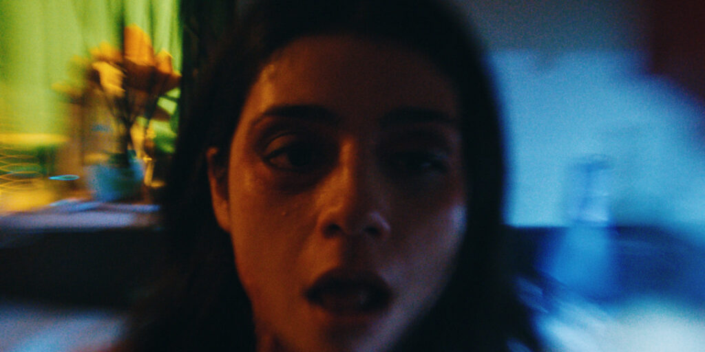 A close-up, slightly blurry image of a woman with dark hair looking into the camera. Her mouth is partly open, and she has a bruise and swelling around her left eye. The background is out of focus with some green and yellow tones.