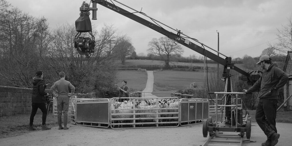 A camera crew films a group of sheep in a pen using a large crane camera on a rural road. Three people stand near the pen, whilst another operates the camera equipment. The scene is set outdoors with trees and fields.