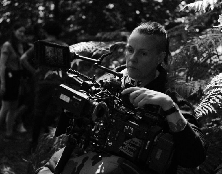 A person operates a large film camera outdoors among ferns and trees, with other people blurred in the background. The scene is in black and white.