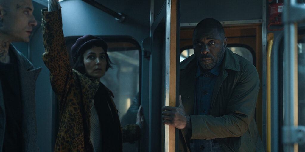 Three people stand inside a dimly lit bus or train. A man in front grips a pole and looks forward; behind him, a woman in a beret and leopard-print coat stands with her hand up. Another man stands to the left.