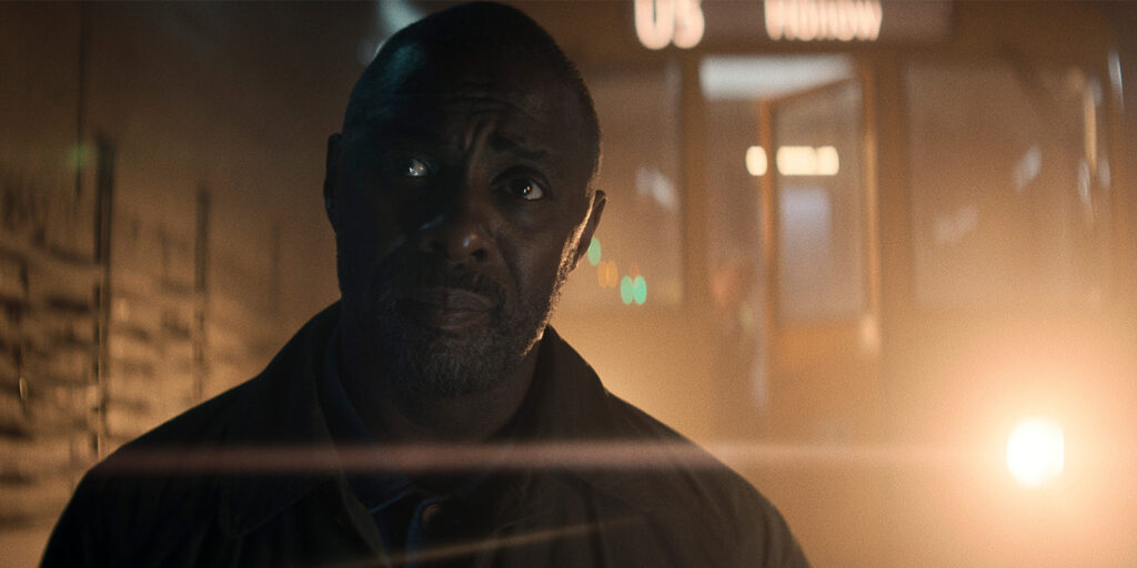 A man with a serious expression stands in a dimly lit area, with light flaring behind him and an open door in the background. The scene appears tense and dramatic.