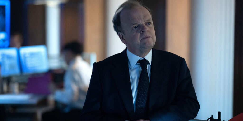 A man in a dark suit and tie stands indoors, looking thoughtful. In the background, a person sits at a desk with computer monitors and papers, suggesting an office or workplace setting.