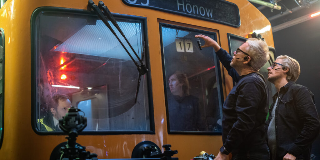 Two people stand outside the window of a yellow train carriage labelled Hönow, with one reaching up towards the glass. Another person is visible inside the train, and camera equipment is set up in the foreground.