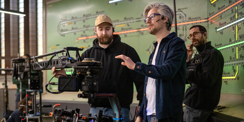 Three people stand next to a camera set-up in front of a large wall diagram. One person operates the camera, another gestures towards it, and the third watches whilst holding a device.