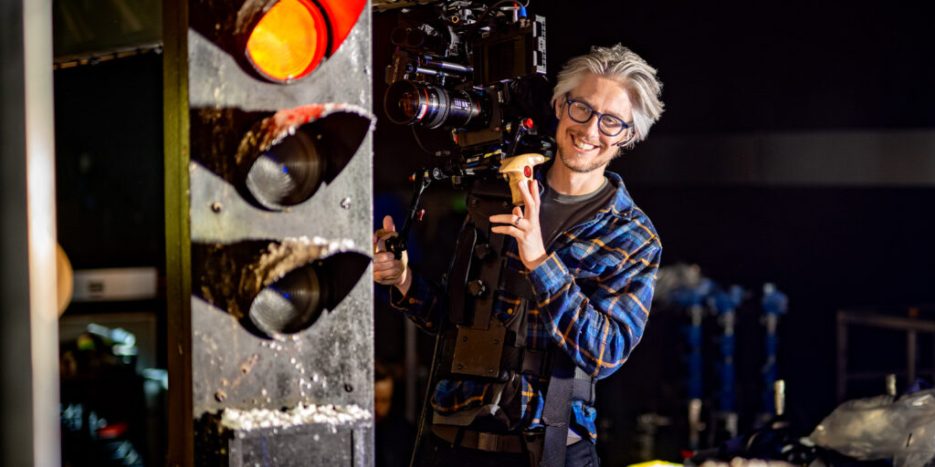 A person wearing glasses and a checked shirt operates a large professional video camera near a traffic light prop in a studio setting.