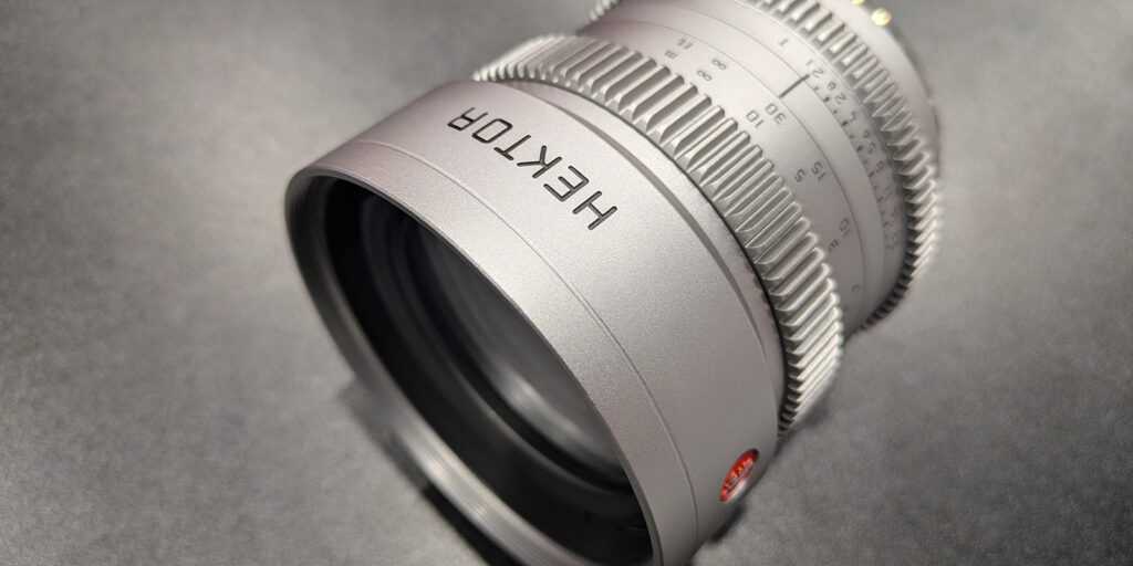 A close-up of a silver camera lens with the brand name HEKTOR engraved on the barrel, showing detailed focus and aperture markings against a plain grey background.