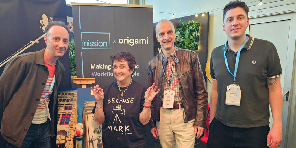 Four people stand indoors in front of a “mission x origami” banner. Three men and one woman face the camera. The woman wears a “Because Mark” T-shirt and the others are dressed casually.