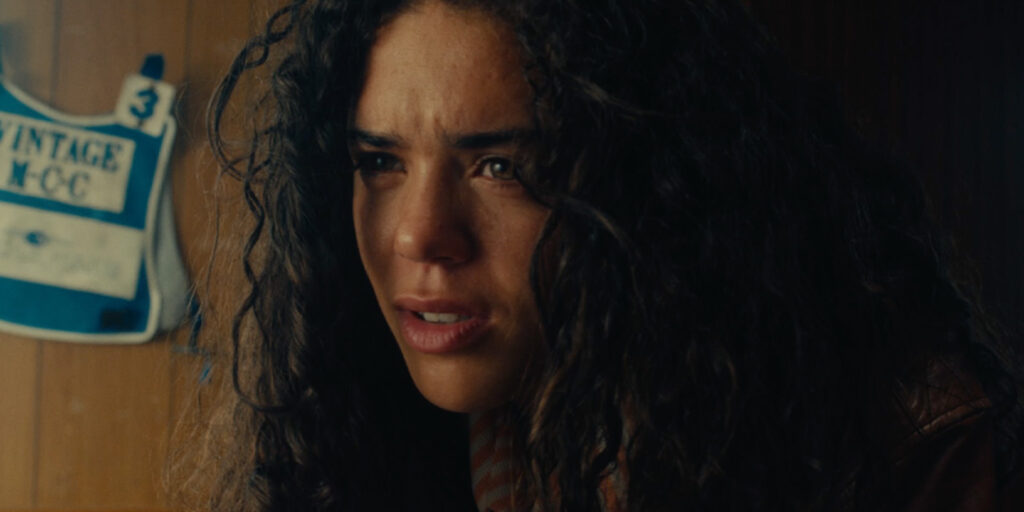 A woman with long, curly hair looks forward with a serious, slightly concerned expression. A sign with the text VINTAGE MCC and the number 3 is visible in the blurry background.