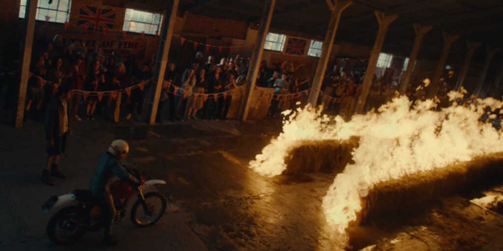 A person on a motorbike prepares to jump over flaming hay bales inside a warehouse, whilst a crowd watches from behind fences decorated with bunting and British flags.