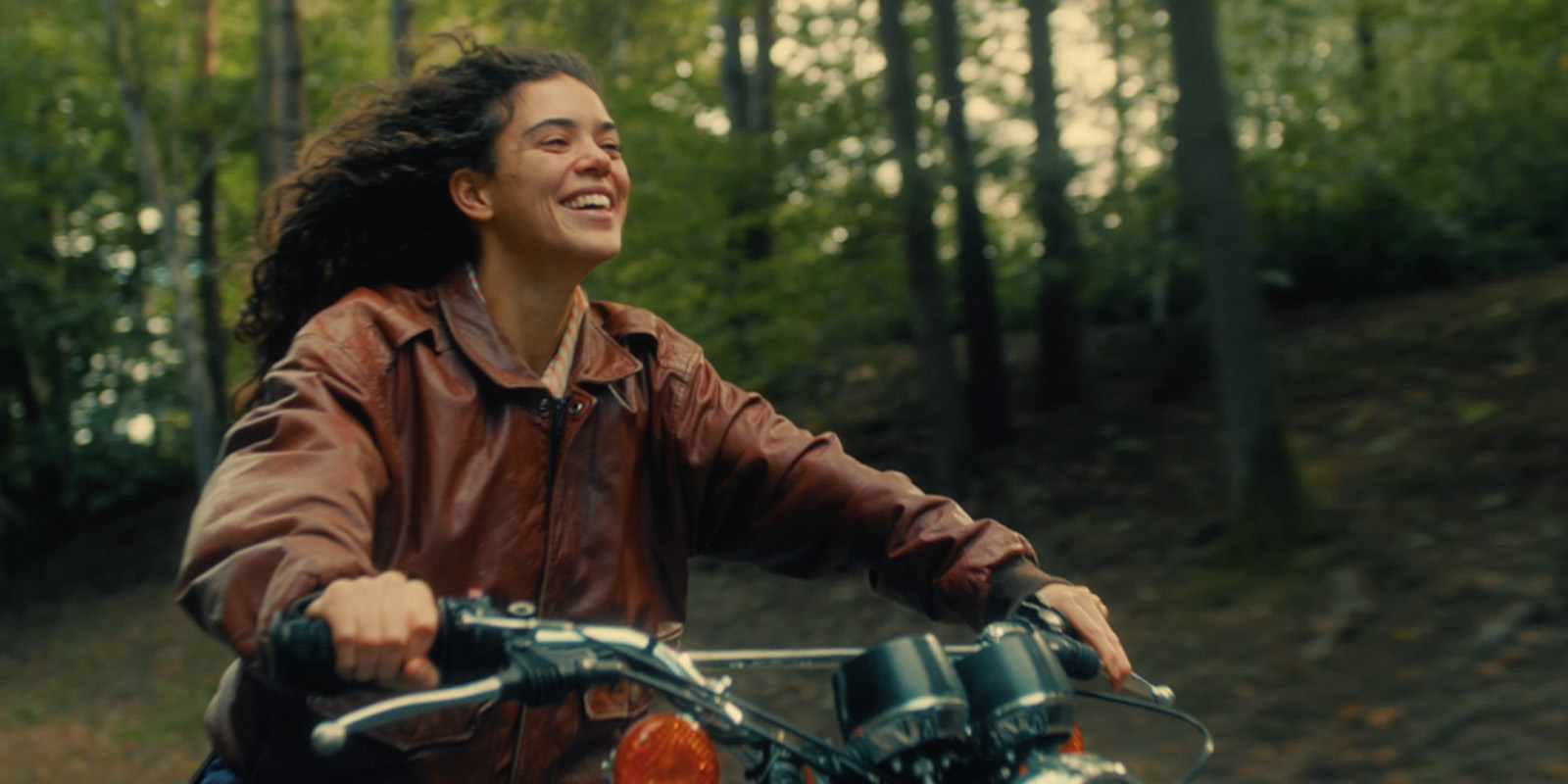 A person with long curly hair wearing a brown leather jacket rides a motorbike through a wooded area, smiling whilst looking ahead.