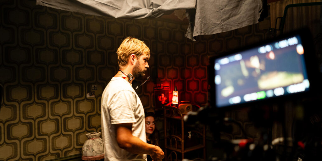 A man in a white shirt stands on a film set with retro-patterned wallpaper, a woman partly visible behind him, and a camera with a blurred screen in the foreground.