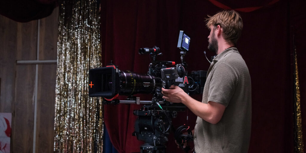 A person operates a professional video camera on a tripod in a studio with gold tinsel and red curtains in the background. The camera has a monitor attached, and the person is focussed on filming.